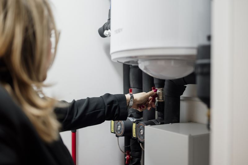 A person is adjusting a valve in a utility room where plumbing equipment is located. This action is part of routine maintenance in a home. Sunlight is present in the space.